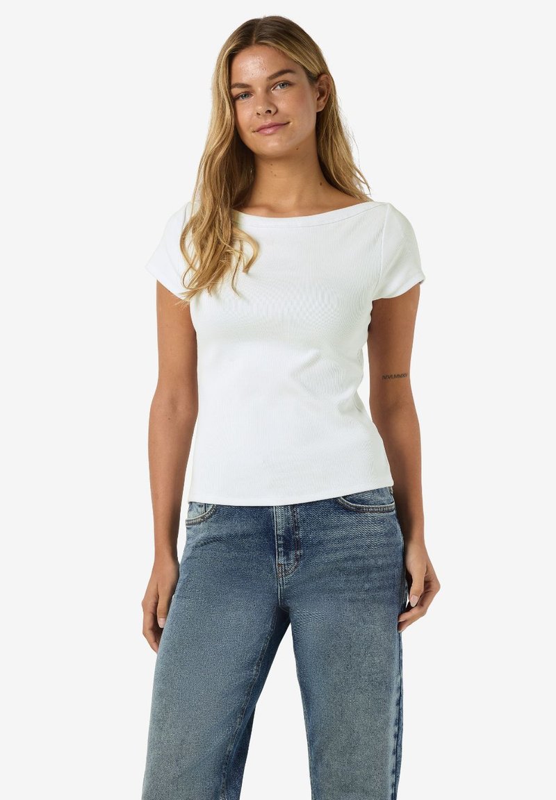 Γυναίκα που στέκεται, φορώντας λευκό τοπ με κοντά μανίκια και μπλε τζιν, με ανοιχτόξανθα χαλαρά μαλλιά και ουδέτερη έκφραση.