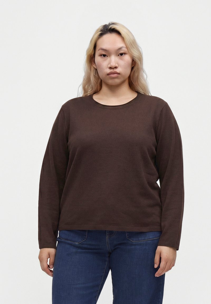 Jeune personne aux longs cheveux blonds porte une chemise marron à manches longues et un jean bleu, debout devant un fond clair uni.