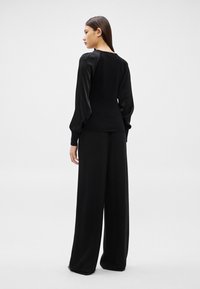 Sieviete ar taisniem tumšiem matiem, tērpusies melnā blūzē ar pūstām piedurknēm un vaļīgās, platas melnas biksēs, skatās prom.