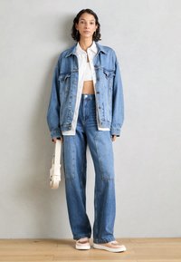 Ljusblå oversize denimjacka, vit croptopp och jeans med raka ben. Modellen håller en krämfärgad väska och bär ljusa sneakers.