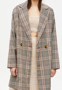 Dubbelknäppt rutigt yllekapp i grå, beige och orange nyanser, med breda slag, två framfickor och oversize-design med lös passform.