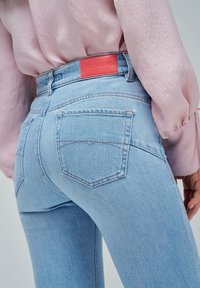 Los vaqueros de denim azul claro presentan un diseño de cintura alta, dos bolsillos traseros con detalles cosidos y una etiqueta roja en la cinturilla.