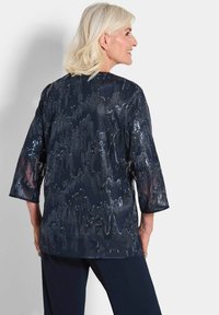 Marineblaue, semi-transparente Bluse mit 3/4-Ärmeln, die ein strukturiertes, schimmerndes Muster und eine glatte Oberfläche aufweist. Kombiniert mit einfarbigen navyblauen Hosen.