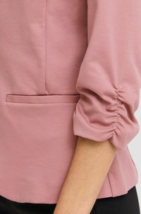 Rózsaszín blúz puha anyagból, összegyűrt ujjakkal és rejtett oldalzsebekkel. Simább textúra, laza szabás.