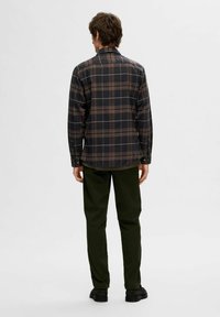 Vista posteriore di un uomo che indossa una camicia a scacchi scuri con quadrati marroni, beige e neri, abbinata a pantaloni verde scuro e scarpe nere.
