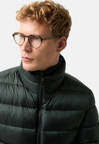 Dunkelgrüne gepolsterte Jacke mit hohem Kragen und Reißverschluss. Der Stoff ist glatt, mit horizontalen Nähten und einem leichten Aussehen.