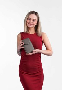 Šedá kožená peněženka s klopou, vybavená zipem a logem. Peněženka je držena osobou v dobře padnoucích červených šatech na bílém pozadí.