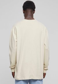 Tricou bej cu mânecă lungă, cu croială lejeră, decolteu rotund și umeri lăsați, având o textură simplă și detalii minime de cusătură.