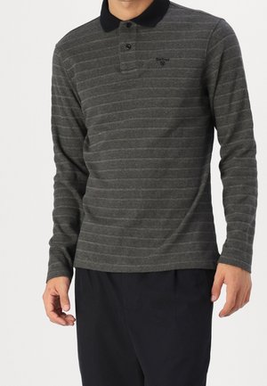 Homme portant un polo à manches longues rayé gris foncé avec un col noir et un pantalon noir, debout devant un fond blanc.