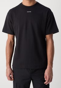 T-shirt en coton noir avec manches courtes et col rond, arborant un logo blanc "Calvin Klein" centré sur la poitrine.