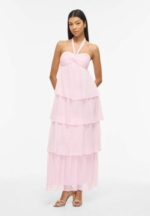 Nő, aki világos rózsaszín, ujjatlan, réteges maxi ruhát visel, pántos nyakrésszel és arany nyitott orrú magassarkúban áll, fehér háttér előtt.