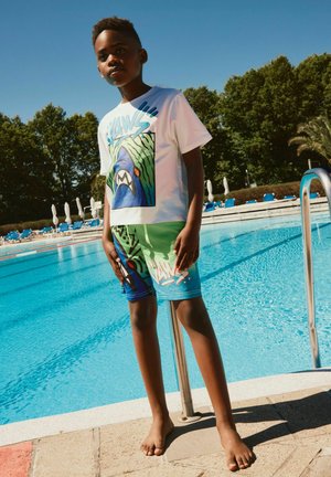Dreng i grafisk t-shirt og shorts står barfodet ved en udendørs swimmingpool på en solrig dag.