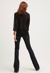 Donna con capelli castani che indossa una blusa nera trasparente e jeans a zampa scuri, in piedi con le spalle alla macchina fotografica su uno sfondo neutro.