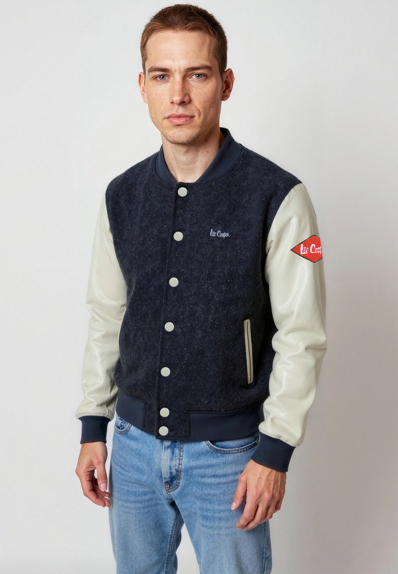 Junger Mann, der eine marineblaue und weiße Lee Cooper Collegejacke mit Knopfleiste und hellblaue Jeans trägt, vor einem schlichten Hintergrund steht.