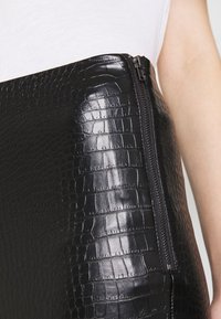 Svart fusk läderkjol med krokodilmönster, med en sidogfång för stängning. Slät, blank yta med distinkta upphöjda mönster.