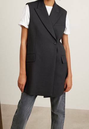 Osoba ubrana w długi czarny bezrękawnik w formie blezera na białą koszulę z krótkim rękawem oraz wyblakłe szare dżinsy, stojąca na jednolitym tle.