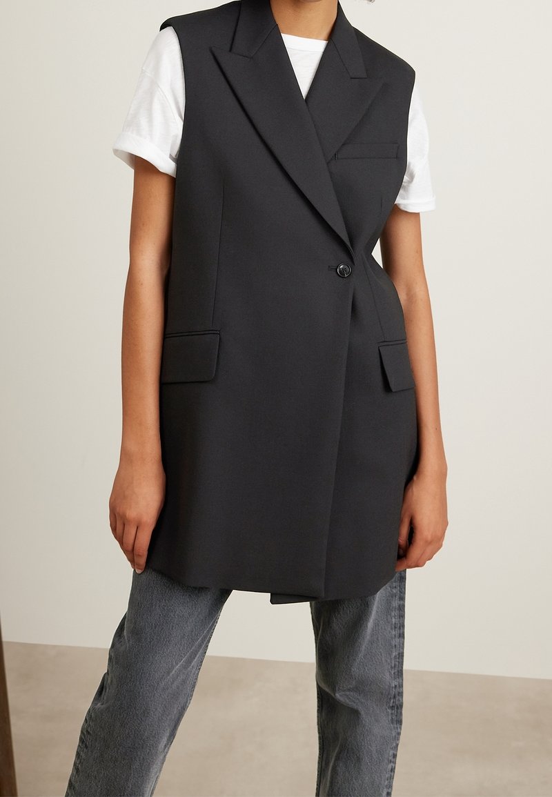 Osoba ubrana w długi czarny bezrękawnik w formie blezera na białą koszulę z krótkim rękawem oraz wyblakłe szare dżinsy, stojąca na jednolitym tle.