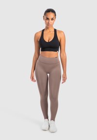 Reggiseno sportivo nero a fascia con schiena scoperta e leggings marroni a vita alta. I leggings hanno una texture liscia. Sono indicate delle scarpe sportive bianche. Sfondo neutro.