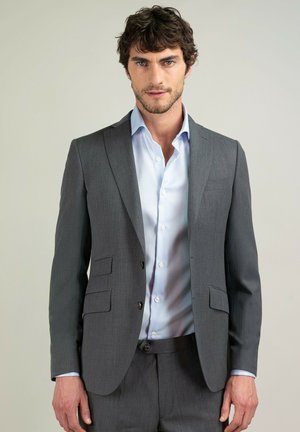 Hombre con chaqueta y pantalones de traje grises y camisa de vestir azul claro, de pie frente a un fondo claro liso.