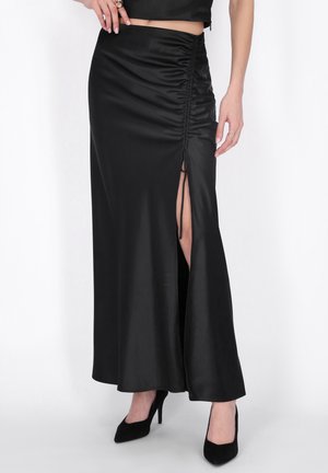 Jupe maxi en satin noir avec détails froncés sur le côté et fente haute sur la cuisse, portée avec des talons hauts noirs et un haut court noir.