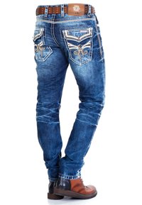 Ausgewaschene blaue Jeans aus Denim mit aufwendigen gestickten Taschen, einem strukturierten braunen Gürtel und braunen Schnürstiefeln mit kontrastierenden Farben.