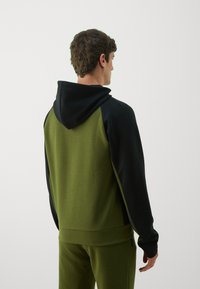Kapuzensweatshirt mit schwarzem Oberteil und olivgrünem Körper, ausgestattet mit einer weichen Textur und Raglanärmeln, kombiniert mit passenden olivgrünen Hosen.