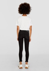 Jeans skinny negros con dobladillo deshilachado, combinados con una camiseta blanca que presenta adornos de ojal en las mangas. El calzado es blanco con suela beige.