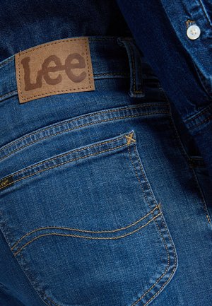 Nærbilde av baklomme på blå denimjeans med brunt lærmerke fra Lee og synlige sømdetaljer.