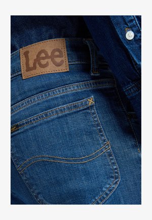 Nærbilde av baklomme på blå denimjeans med brunt lærmerke fra Lee og synlige sømdetaljer.