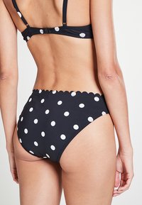 Bas de bikini noir taille haute avec des pois blancs, ceinture à bord festonné, texture lisse et design couvrant à l'arrière.