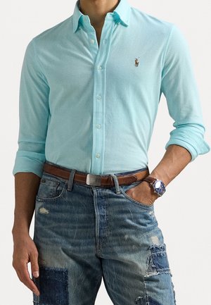 Man som bär en ljusblå skjorta med knäppning, brunt läderbälte, slitna lapptäckesjeans och en armbandsklocka med blå urtavla på höger handled.