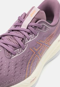 Zapatilla deportiva morada con parte superior de malla y acentos naranjas. Cuenta con cordones, collar acolchado y suela texturizada. Diseño ligero, forma aerodinámica.