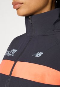 Juodas zip-up švarkas su ryškiai oranžine juosta per krūtinę ir pilka logo. Sklandi tekstūra su aukštu apykakle.