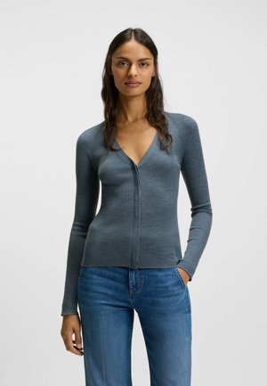 Cardigan a coste, con bottoni, di un morbido colore blu-grigio, caratterizzato da un profondo scollo a V e maniche lunghe, abbinato a jeans blu attillati.