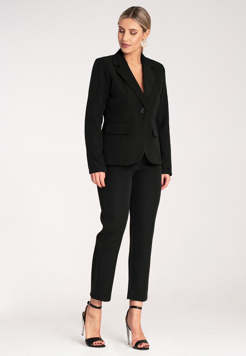 Czarna taliowana marynarka z pojedynczym guzikiem, dwoma przednimi kieszeniami, w połączeniu z dopasowanymi czarnymi spodniami i sandałami na obcasie z paskami. Gładki materiał.