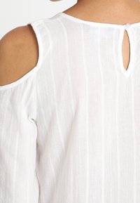 Blusa bianca con un motivo a righe testurizzato, con ritagli sulle spalle e chiusura a bottoni sul retro. Tessuto morbido e leggero.