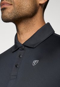 Polo noir en tissu doux, avec un plastron à deux boutons et un logo brodé sur la poitrine gauche. Design de col lisse.