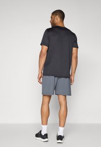 Man som står med ryggen mot kameran, iförd en svart t-shirt med korta ärmar, grå shorts, vita strumpor och svarta sportskor mot en enfärgad bakgrund.