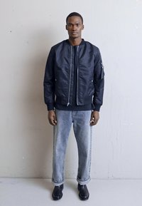 Bomberjakke i sort nylon med en frontlynlås, ribbede manchetter og sidelommer, parret med lyse denimjeans og sorte sko.