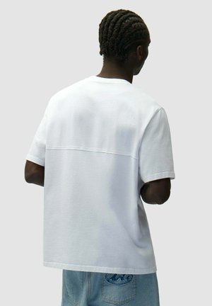 Uomo con capelli intrecciati visto di schiena, che indossa una t-shirt bianca a maniche corte semplici e jeans di denim azzurro chiaro con una toppa sulla tasca posteriore.