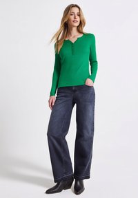 Zielony top z długim rękawem z okrągłym dekoltem i plisą zapinaną na trzy guziki, zestawiony z ciemnoniebieskimi szerokimi spodniami jeansowymi oraz czarnymi botkami na koturnie.