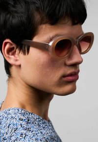 Överdimensionerade, rundade solglasögon med en genomskinlig ljusbrun ram och mörka tonade linser. Texturerad bakgrund, modell som bär en blå och grå stickad tröja.