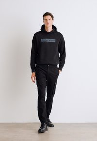 Hanorac negru cu glugă, având un logo albastru Calvin Klein. Asortat cu pantaloni cargo negri și pantofi negri. Culori solide, design casual.