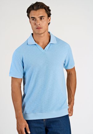 Uomo con polo a maniche corte azzurro chiaro e jeans blu scuro, in piedi davanti a uno sfondo bianco semplice.