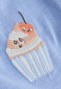 Gesticktes Cupcake-Design mit weißer Glasur, pfirsichfarbenen Schichten und einer Kirsche oben drauf, auf einem hellblauen Stoffhintergrund.