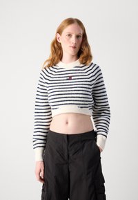 Kunststrikket genser med maritime blå- og hvite striper, rund hals, og liten brodert logo på brystet. Kombinert med mørke bukser.