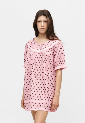 Jeune femme portant une robe rose à manches courtes avec des pois marron, debout devant un fond gris clair uni.