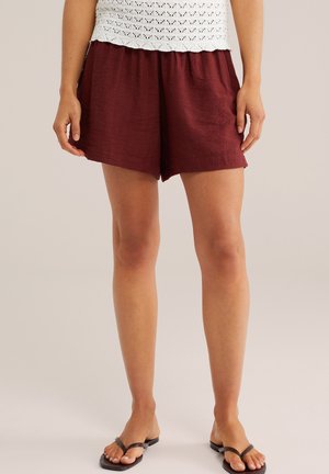 Kobieta w luźnych, ciemnoczerwonych szortach, białym szydełkowanym topie i czarnych klapkach stoi na jednolitym tle.