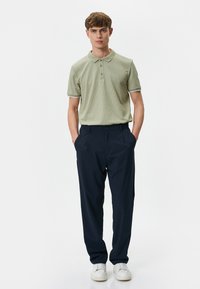 Lys grøn polo shirt med en tekstureret overflade og hvid kant, parret med marineblå bukser og hvide sneakers. Modellen står foran en neutral baggrund.