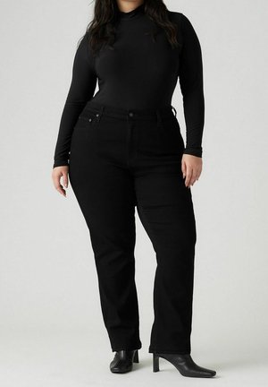 Kvinde står iført sort tætsiddende langærmet top, sorte straight-leg jeans og sorte ankelstøvler med hæl mod neutral baggrund.